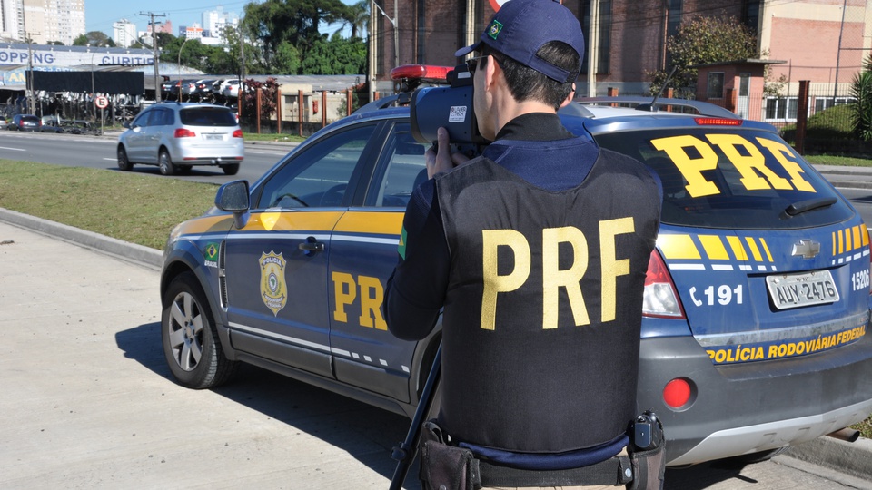 concurso prf: a imagem mostra policial rodoviário segurando equipamento portátil que mede velocidade ao lado de viatura da PRF