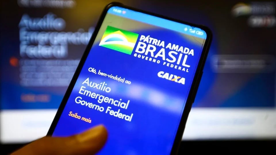 Contestar auxílio emergencial 2021: mão segurando celular. Na tela do aparelho, é possível ver a página do programa auxílio emergencial